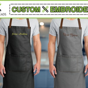 May include: Two people wearing aprons with custom embroidery. The apron on the left is black with yellow embroidery that says "Jerry's Cooking". The apron on the right is gray with brown embroidery that says "Your Name".
