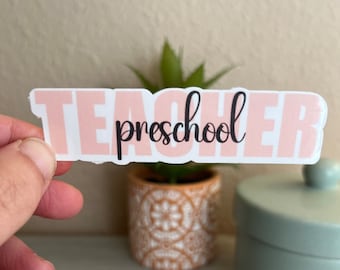 May include: A pink and white sticker with the text "Teacher preschool" in black lettering.