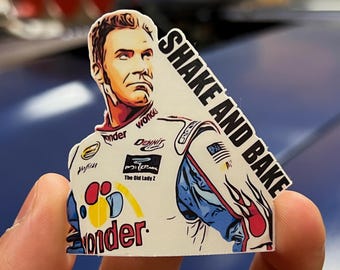 May include: A white sticker with a cartoon illustration of a race car driver wearing a blue and red racing suit. The driver is holding a steering wheel and the sticker says "Shake and Bake".