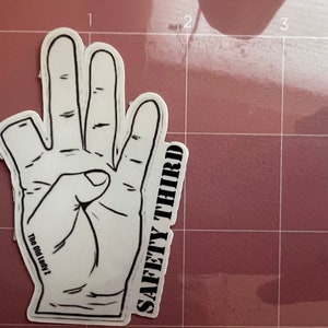 Pu&ograve; includere: Un adesivo bianco con un contorno nero di una mano che fa il gesto di 'sicurezza al terzo posto'. L'adesivo ha il testo 'SAFETY THIRD' scritto verticalmente sul lato destro e 'The Old Lady 1' scritto orizzontalmente sul lato sinistro.