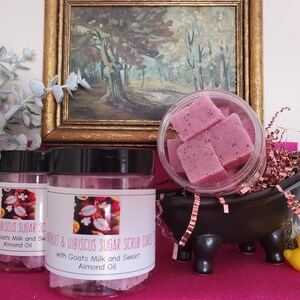 May include: Fruit & Hibiscus Sugar Scrub Cubes with Goat's Milk and Sweet Almond Oil. The image shows two clear jars with black lids and labels. A small black bathtub holds pink cubes. A yellow rubber duck is also present.