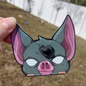 Puede incluir: Una pegatina con la imagen de una cabeza de murciélago estilizada con orejas grandes y una marca en forma de corazón. El murciélago tiene un cuerpo gris, orejas y boca rosas y ojos blancos. La pegatina se sostiene sobre un fondo de hierba.