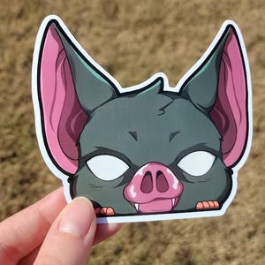 May include: A sticker featuring a cartoon bat face with large ears and a pink nose and ears. The bat has white eyes and is outlined in black and white. The sticker is held against a blurred background of dry grass.