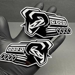 Puede incluir: Dos pegatinas troqueladas en blanco y negro con un diseño de motor estilizado. Las pegatinas tienen un estilo gráfico audaz con un contorno blanco y se sostienen en una mano enguantada. El diseño incluye una cabeza de carnero y tubos de escape.