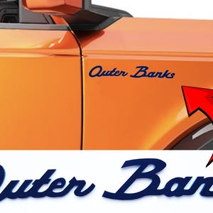 May include: Orange vehicle panel with the text "Outer Banks" in a dark blue, cursive font. A red arrow points to the text. The lower portion of the image shows the same text in a larger size.