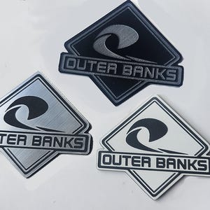 Puede incluir: Tres pegatinas con el logotipo de Outer Banks en forma de diamante. Las pegatinas presentan un diseño de ola y las palabras "Outer Banks". Una pegatina es negra, otra plateada y otra blanca.