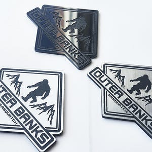 Puede incluir: Tres insignias metálicas de Outer Banks Sasquatch. Cada insignia presenta una silueta de un Sasquatch, montañas y las palabras "Outer Banks Sasquatch". Las insignias son en negro, plata y una combinación de negro y blanco.