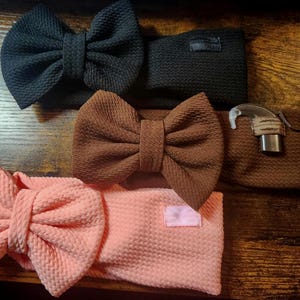 May include: Three textured headbands with large fabric bows in black, brown, and light pink. The brown headband features a small attached hearing aid device with a clear ear hook and a metallic component. Each band has a rectangular fabric tag.
