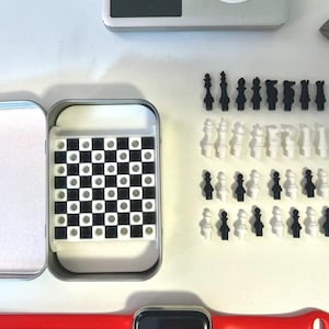 Pode incluir: Um conjunto de xadrez de viagem de metal prateado com um tabuleiro de xadrez preto e branco e pe&ccedil;as de xadrez pretas e brancas. O conjunto est&aacute; aberto e as pe&ccedil;as est&atilde;o dispostas sobre uma superf&iacute;cie branca.