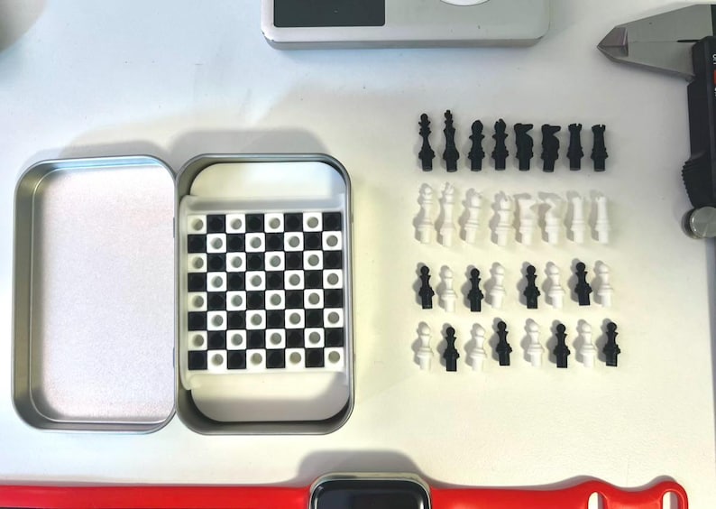 Pode incluir: Um conjunto de xadrez de viagem de metal prateado com um tabuleiro de xadrez preto e branco e pe&ccedil;as de xadrez pretas e brancas. O conjunto est&aacute; aberto e as pe&ccedil;as est&atilde;o dispostas sobre uma superf&iacute;cie branca.