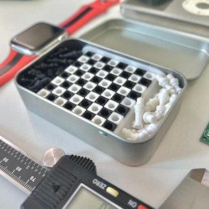 Pode incluir: Uma lata de metal prateada com um design de tabuleiro de xadrez preto e branco no interior. A lata cont&eacute;m pe&ccedil;as de xadrez de pl&aacute;stico branco e um conjunto de xadrez de pl&aacute;stico preto.
