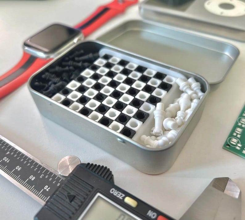 Pode incluir: Uma lata de metal prateada com um design de tabuleiro de xadrez preto e branco no interior. A lata cont&eacute;m pe&ccedil;as de xadrez de pl&aacute;stico branco e um conjunto de xadrez de pl&aacute;stico preto.