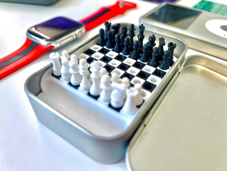 Pode incluir: Um pequeno conjunto de xadrez port&aacute;til em uma lata de metal prateada. As pe&ccedil;as de xadrez s&atilde;o pretas e brancas e est&atilde;o dispostas em um tabuleiro de xadrez preto e branco. A lata est&aacute; aberta e as pe&ccedil;as de xadrez s&atilde;o vis&iacute;veis.