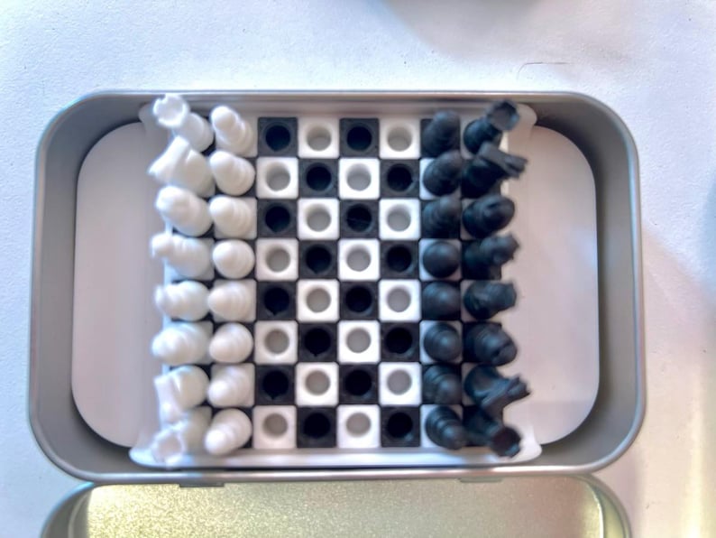 Pode incluir: Uma lata prateada com um design de tabuleiro de xadrez branco e preto. O tabuleiro de xadrez tem quadrados pretos e brancos. H&aacute; pe&ccedil;as de xadrez brancas e pretas no tabuleiro.