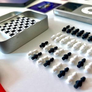 Pode incluir: Um conjunto de pe&ccedil;as de xadrez pretas e brancas sobre uma superf&iacute;cie branca. As pe&ccedil;as s&atilde;o feitas de pl&aacute;stico e t&ecirc;m cerca de 2,5 cm de altura. H&aacute; tamb&eacute;m uma pequena caixa de metal com um tabuleiro de xadrez dentro.