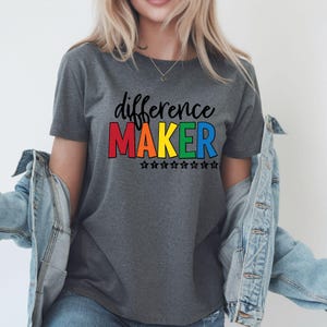 Op de afbeelding: Een grijs T-shirt met de tekst "difference maker" in regenboogkleuren. De tekst is in een speels lettertype en wordt omringd door kleine zwarte sterren.
