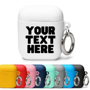 May include: A white Airpods case with a black text overlay that says "YOUR TEXT HERE". The case has a silver metal ring attached to it. The image also shows a variety of colored Airpods cases in yellow, red, pink, blue, turquoise, gray, light blue, and black.