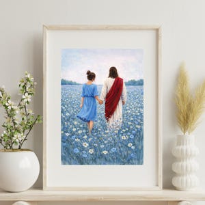 May include: Framed art print depicting two figures walking through a field of blue flowers. One figure wears a blue dress, the other a white robe with a red sash. The artwork is displayed in a light-colored wooden frame.