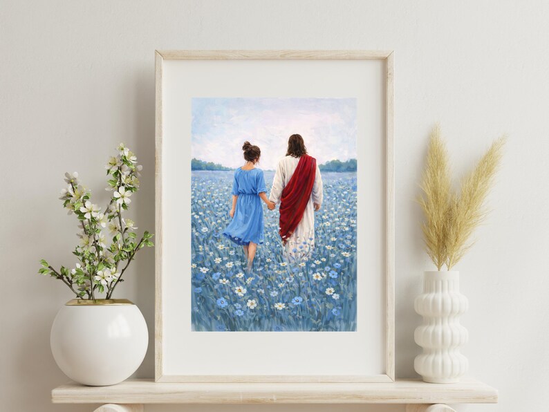 May include: Framed art print depicting two figures walking through a field of blue flowers. One figure wears a blue dress, the other a white robe with a red sash. The artwork is displayed in a light-colored wooden frame.