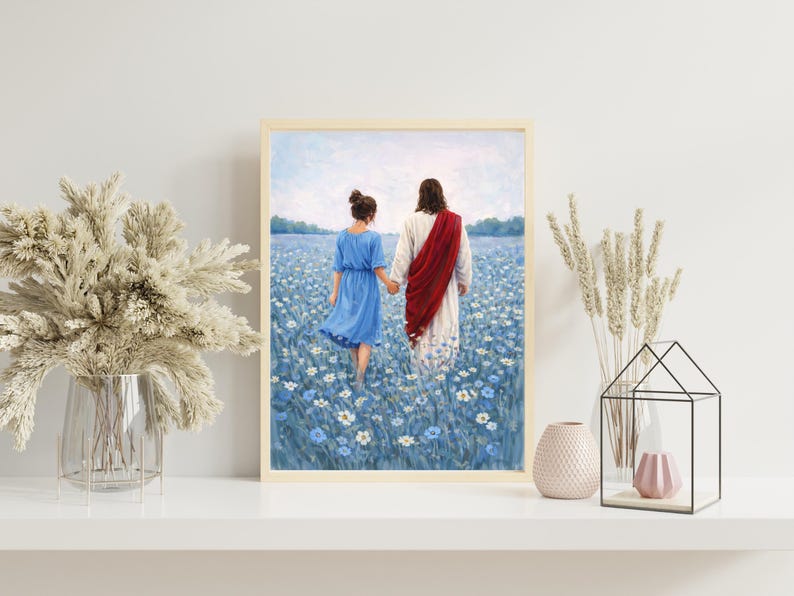 May include: Framed art print depicting two figures walking through a field of blue flowers. One figure wears a blue dress, the other a white robe and red sash. The artwork is displayed on a white shelf with decorative elements.
