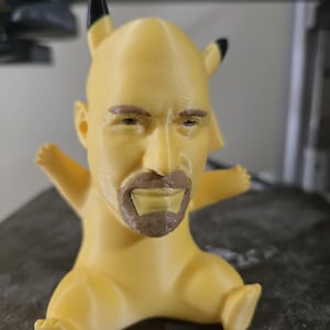 Può includere: Una statuetta di Pikachu stampata in 3D di colore giallo con il viso di Dwayne "The Rock" Johnson. La statuetta ha le orecchie nere e una barba marrone.