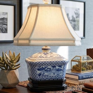 May include: A decorative table lamp with a cream-colored lampshade and a blue and white porcelain base. The base features a landscape design. The lamp sits on a dark wooden table with other decorative items.