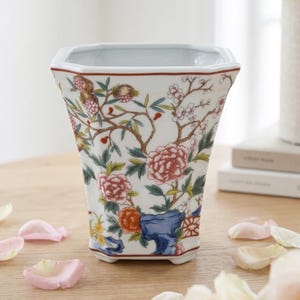 May include: A white ceramic vase with a square top and a floral design. The vase features pink, red, blue, and green flowers and branches. The vase is approximately 8 inches tall.