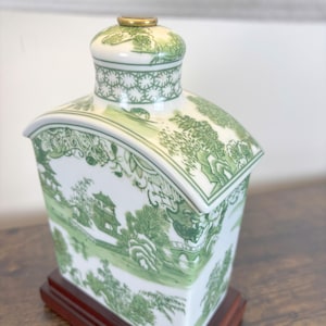 May include: A white and green ceramic tea caddy with a landscape design, featuring pagodas and trees. The caddy has a rectangular shape with a curved top and sits on a dark wooden base. The lid is topped with a gold-coloured accent.