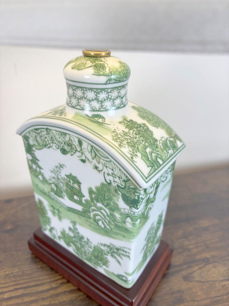 May include: A white and green ceramic tea caddy with a landscape design, featuring pagodas and trees. The caddy has a rectangular shape with a curved top and sits on a dark wooden base. The lid is topped with a gold-coloured accent.