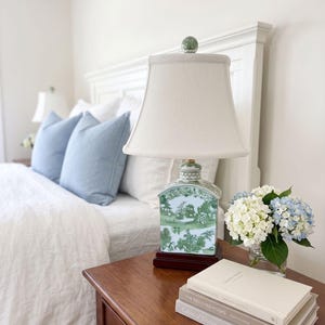 May include: A bedroom scene with a wooden bedside table holding a lamp, books, and flowers. The lamp has a white shade and a green and white ceramic base. Blue cushions and white bedding are on the bed.