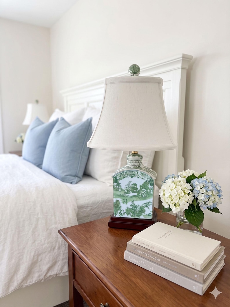 May include: A bedroom scene with a wooden bedside table holding a lamp, books, and flowers. The lamp has a white shade and a green and white ceramic base. Blue cushions and white bedding are on the bed.
