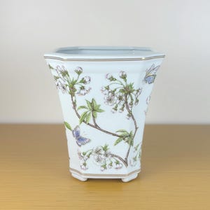 May include: White ceramic planter with a floral design featuring cherry blossoms and butterflies. The planter has a hexagonal shape and a gold rim.