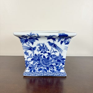 May include: A white porcelain vase with a square shape and flared sides. The vase is decorated with blue floral and bird motifs. A Greek key pattern borders the base. The vase is a decorative item.