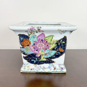 May include: A white square planter with a floral design. The design features large blue leaves, pink and yellow flowers, and gold accents.