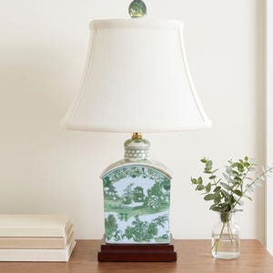 May include: A ceramic table lamp with a white lampshade and a green and white landscape design. The lamp sits on a dark brown wooden base. A small glass vase with greenery is on the table.
