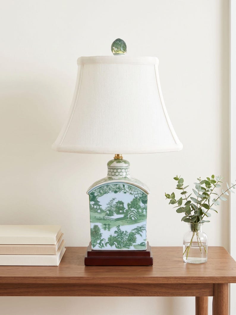 May include: A ceramic table lamp with a white lampshade and a green and white landscape design. The lamp sits on a dark brown wooden base. A small glass vase with greenery is on the table.