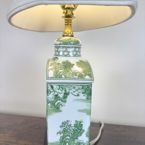 May include: A ceramic table lamp with a white lampshade. The lamp base is rectangular with a green and white landscape design. The lamp has a brass-coloured neck and a white cord.