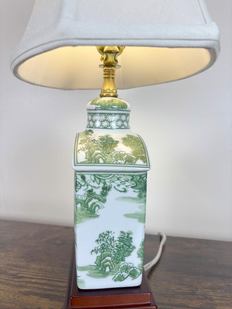 May include: A ceramic table lamp with a white lampshade. The lamp base is rectangular with a green and white landscape design. The lamp has a brass-coloured neck and a white cord.