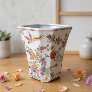 May include: A white, hexagonal ceramic vase with a colorful floral and bird design. The vase features a red rim and is decorated with orange, blue, and green flowers and a bird. The vase is sitting on a wooden surface.