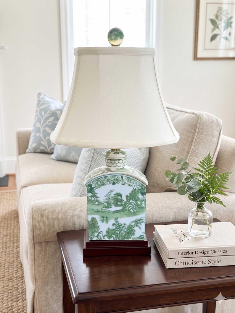 May include: A decorative table lamp with a white lampshade and a green and white porcelain base featuring a landscape design. The lamp sits on a dark wood base and is topped with a glass orb finial. The lamp is on a dark wood table.