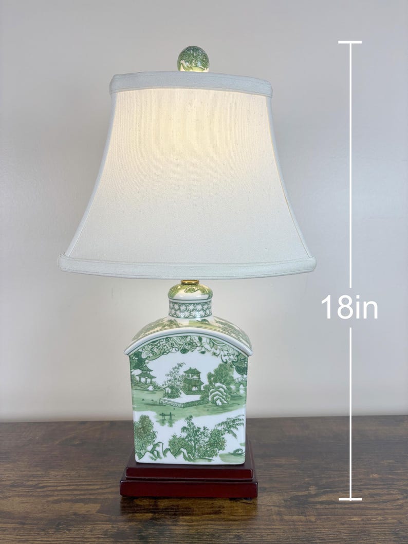 May include: A table lamp, approximately 46 cm tall, with a white lampshade and a ceramic base. The base has a dark red wooden stand and a green and white landscape design. The lamp is topped with a decorative glass orb.