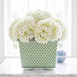 May include: A rectangular ceramic planter with a green and white geometric pattern, filled with white hydrangea flowers. The planter sits on small white feet. A small blue and white decorative jar is in the background.