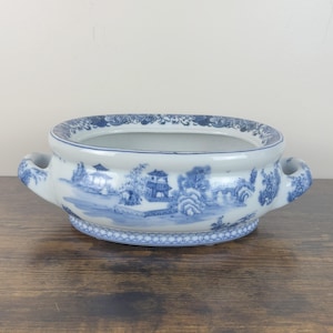 May include: A blue and white porcelain planter with a landscape design. The rectangular planter has two curved handles and a detailed pattern around the rim and base. The landscape scene depicts buildings, trees, and bridges.