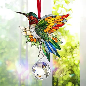 May include: A stained glass hummingbird suncatcher with a crystal prism hanging from it. The hummingbird is depicted with vibrant colors, including green, blue, yellow, and red. The suncatcher is surrounded by flowers and leaves.