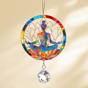 May include: A stained glass suncatcher featuring a colorful depiction of a person in a yoga pose sitting on a lotus flower. The suncatcher is suspended from a string and has a crystal prism hanging from the bottom.