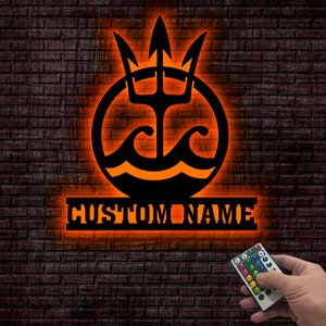 May include: A black metal wall decor with a trident and waves inside a circle, with the words "CUSTOM NAME" below. The decor is illuminated with orange light and a remote control is shown in the bottom right corner.