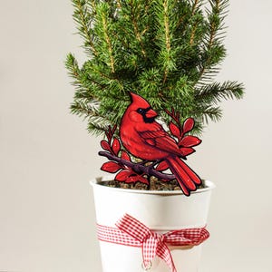 May include: A red cardinal bird figurine perched on a branch with red leaves, placed in a white pot with a red and white checkered ribbon. The pot contains a small green fir tree.
