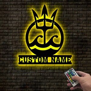May include: A black metal wall decor with a trident and waves inside a circle, with the words "CUSTOM NAME" below. The decor is illuminated with yellow light and a remote control is shown in the bottom right corner.