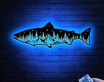 May include: A black metal silhouette of a fish with a mountain and forest scene inside. The fish is lit up with blue light and has a remote control to change the color of the light.