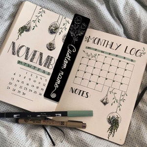 May include: A black bookmark with the text "Custom name" written in white cursive. The bookmark is decorated with a simple floral design. The bookmark is placed on top of a monthly calendar page with the month of November written in cursive. The calendar page is decorated with drawings of hanging plants.